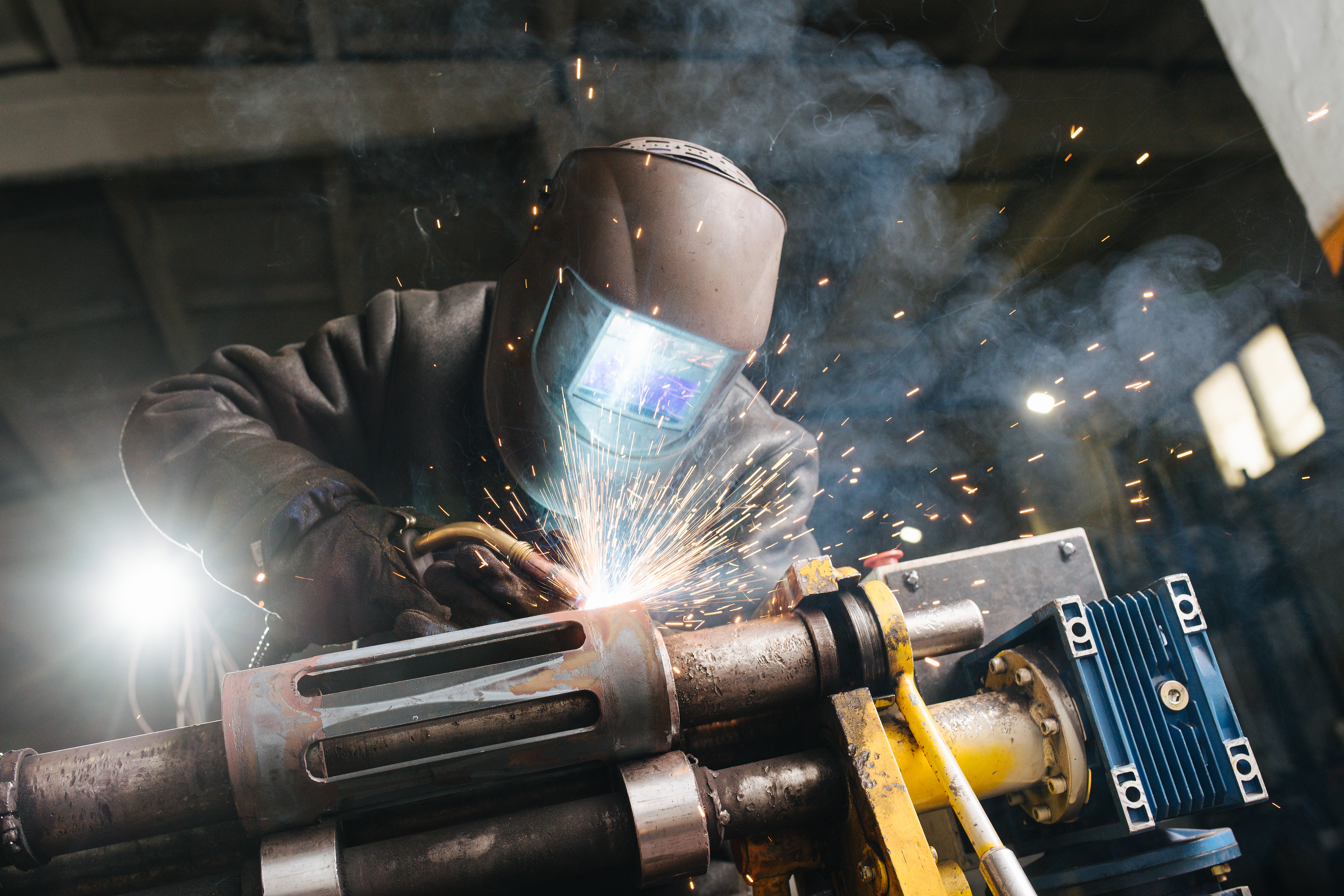 Working welder in protective mask and clothing. He's welding metal piece, fixed with industrial clamp. His mask glowing blue, sparks flying in the air. Workshop is big and resembles hangar. Low angle.